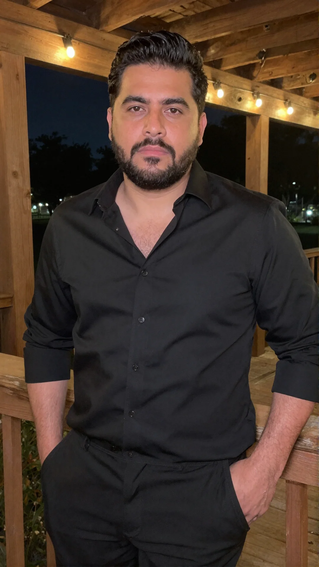 Serious Mexican man in an open-collar black shirt on a back porch with string lights