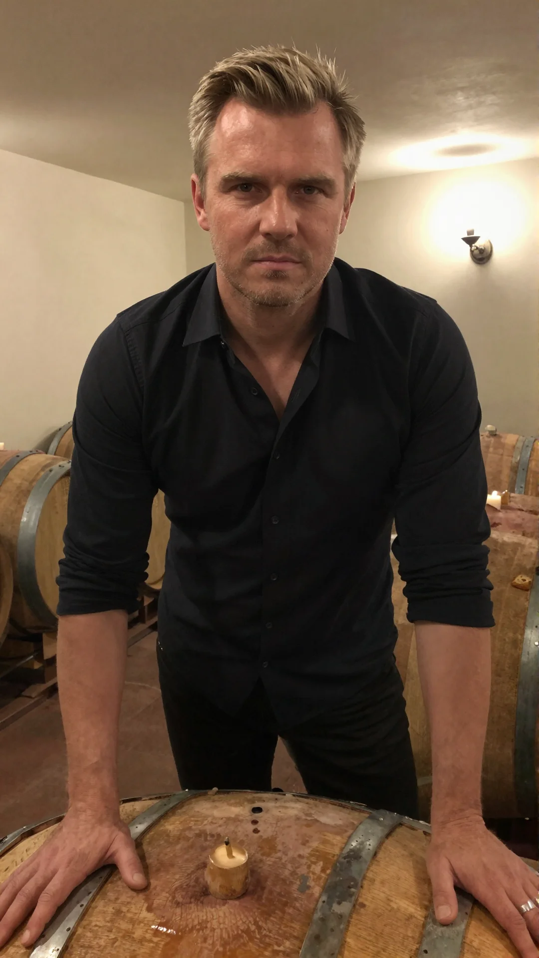 Serious Russian man in an open black shirt leaning on a barrel in a candlelit wine cellar