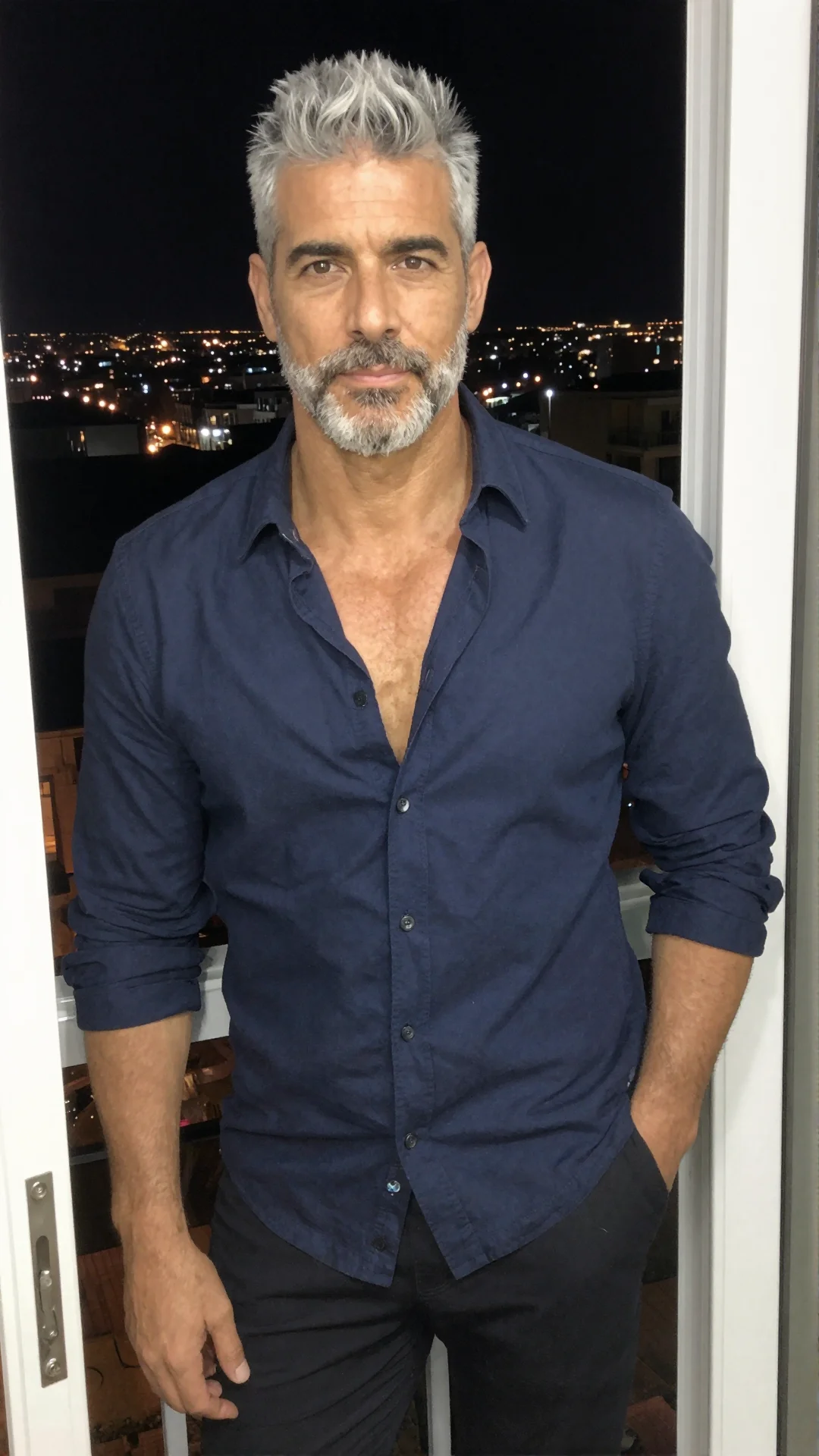Serious Spanish man in an open navy shirt on a balcony doorway at night